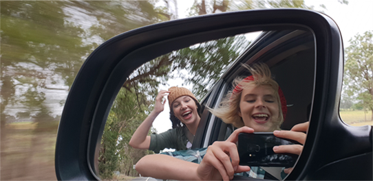 Two people taking their picture in a car side mirror
